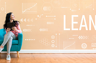 Das Bild zeigt eine Person, die auf einem türkisfarbenen Stuhl sitzt und ein Tablet in den Händen hält. Im Hintergrund befindet sich eine hell pfirsichfarbene Wand, auf der weiße grafische Elemente wie Diagramme, Pfeile und Sechsecke zu sehen sind. Besonders auffällig ist das große weiße Wort „LEAN“, das zentral auf der Wand steht.