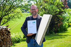 Robert Schwemmer- Green Product Award Dieses Bild zeigt Herrn Robert Schwemmer mit der Urkunde des Green Product Award in der rechten hand und einer Schallschutzplatte aus Hanf in der Linken Hand im Hintergrund findet man einen grünen Garten mit einem Holzstoß