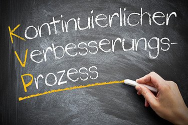 Das Bild zeigt eine schwarze Tafel, auf der in weißer Kreideschrift die deutschen Wörter „Kontinuierlicher Verbesserungsprozess“ geschrieben stehen. Die Anfangsbuchstaben K, V und P sind gelb hervorgehoben und bilden das Akronym „KVP“. Eine Hand hält ein Stück Kreide und zieht eine gelbe Linie unter dem Wort „Prozess“.