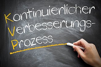 Hanf Das Bild zeigt eine schwarze Tafel, auf der in weißer Kreideschrift die deutschen Wörter „Kontinuierlicher Verbesserungsprozess“ geschrieben stehen. Die Anfangsbuchstaben K, V und P sind gelb hervorgehoben und bilden das Akronym „KVP“. Eine Hand hält ein Stück Kreide und zieht eine gelbe Linie unter dem Wort „Prozess“.