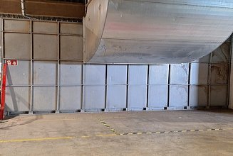 Das Bild zeigt eine Lager- oder Industriehalle mit einem großen Metall-Lüftungsrohr, das an Decke und Wand befestigt ist. Links befindet sich ein Feuerlöscher sowie verschiedene Baumaterialien und Geräte. Rechts sind mehrere Sicherheitsschilder an der Wand angebracht. Der Boden ist mit gelb-schwarzem Warnband markiert, das einen bestimmten Bereich abgrenzt.