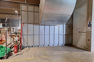 Pausenraum Das Bild zeigt eine Lager- oder Industriehalle mit einem großen Metall-Lüftungsrohr, das an Decke und Wand befestigt ist. Links befindet sich ein Feuerlöscher sowie verschiedene Baumaterialien und Geräte. Rechts sind mehrere Sicherheitsschilder an der Wand angebracht. Der Boden ist mit gelb-schwarzem Warnband markiert, das einen bestimmten Bereich abgrenzt.