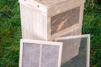 Bienenstock Sumsi Zu Sehen ist ein Bienenstock der mit Hanfplatten gedämmt wurde auf einer grünen Wiese Davor stehen die Innenteile des Bienenstocks, damit die Hanfplatten besser sichtbar sind