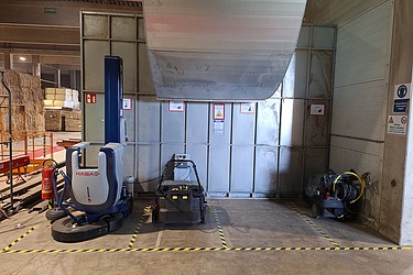 Das Bild zeigt eine Produktionshalle mit mehreren Maschinen. Links steht eine große blau-weiße Maschine mit der Aufschrift „HABASIT“, in der Mitte eine kleinere schwarze Anlage, und rechts ein Gerät mit mehreren Kabeln oder Schläuchen. Der Boden ist mit gelb-schwarzen Sicherheitsmarkierungen versehen. An der Wand hängen Warn- und Hinweisschilder, links ist ein Feuerlöscher zu sehen, und von der Decke verläuft ein großes Metallrohr.