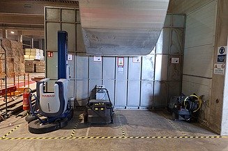 Ladestation Das Bild zeigt eine Produktionshalle mit mehreren Maschinen. Links steht eine große blau-weiße Maschine mit der Aufschrift „HABASIT“, in der Mitte eine kleinere schwarze Anlage, und rechts ein Gerät mit mehreren Kabeln oder Schläuchen. Der Boden ist mit gelb-schwarzen Sicherheitsmarkierungen versehen. An der Wand hängen Warn- und Hinweisschilder, links ist ein Feuerlöscher zu sehen, und von der Decke verläuft ein großes Metallrohr.